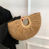 A woven rattan half-moon beach handbag held by a person against a white wall.