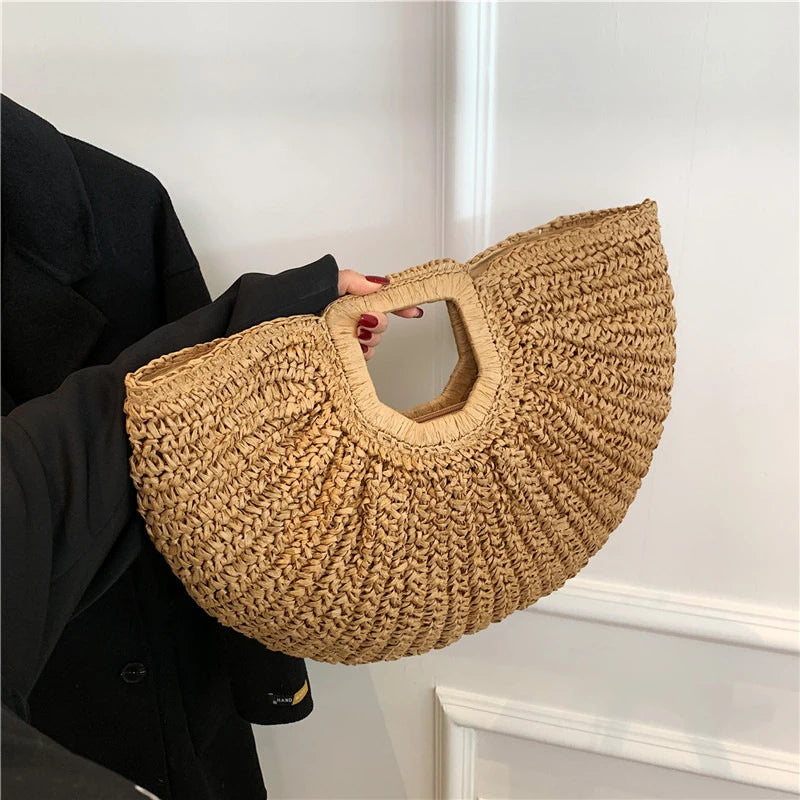 A woven rattan half-moon beach handbag held by a person against a white wall.