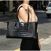 A woman holds a black crocodile embossed leather tote bag with a magnetic closure outdoors.