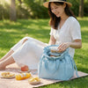 A woman sits on the grass holding a light blue insulated drawstring lunch bag filled with snacks and crackers.