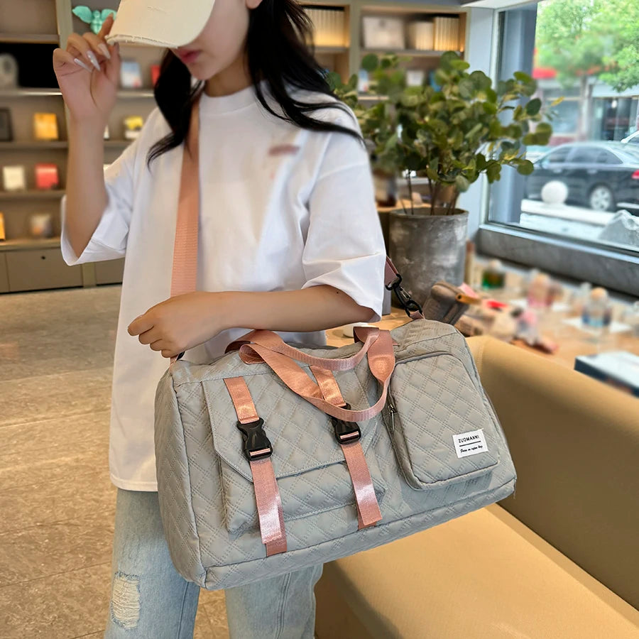 A woman holds a light gray nylon travel duffle bag with an argyle pattern in a casual indoor setting.