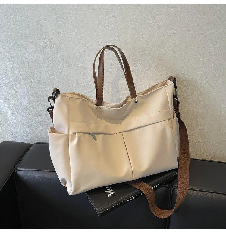 A beige lightweight nylon tote bag with brown leather handles and multiple pockets resting on a black surface.