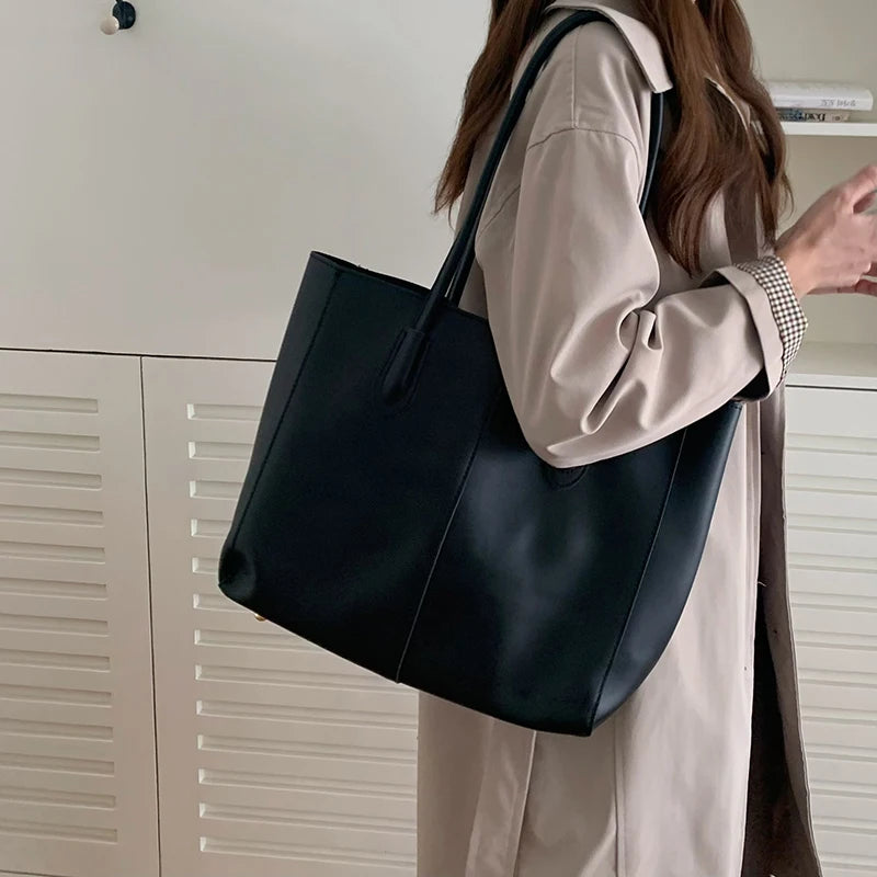 A woman is holding a large black leather tote bag with a shoulder strap, next to white cabinetry.