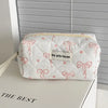A white quilted cosmetic bag with pink ribbon embroidery rests on a white surface.