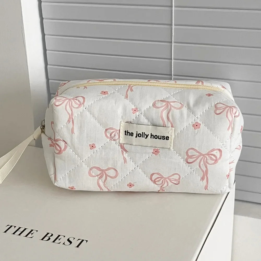 A white quilted cosmetic bag with pink ribbon embroidery rests on a white surface.