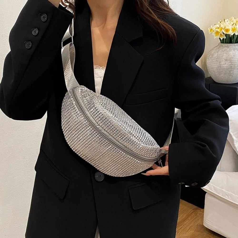 A woman holds a silver rhinestone-embellished crossbody waist bag with an adjustable strap against a black blazer.