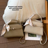A beige PU leather crossbody clutch bag with a satin bow detail resting on a wooden floor next to a white pillow.