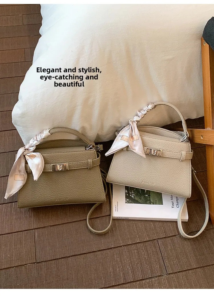A beige PU leather crossbody clutch bag with a satin bow detail resting on a wooden floor next to a white pillow.