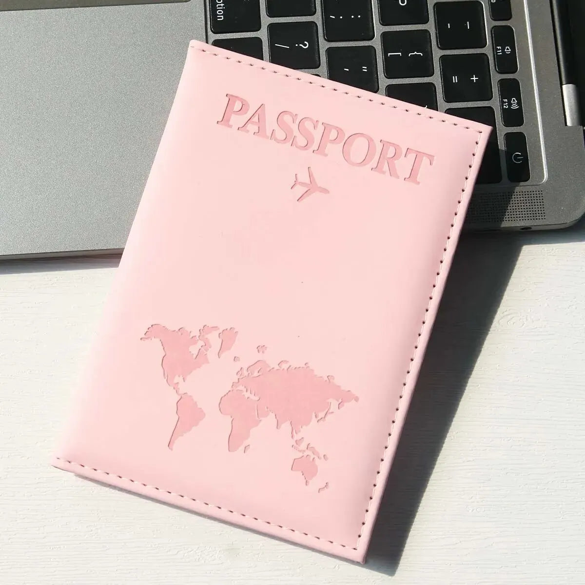 A pink passport holder with a world map design resting on a white surface next to a laptop keyboard.