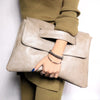 A hand holding a beige leather crossbody clutch bag against a green sweater.