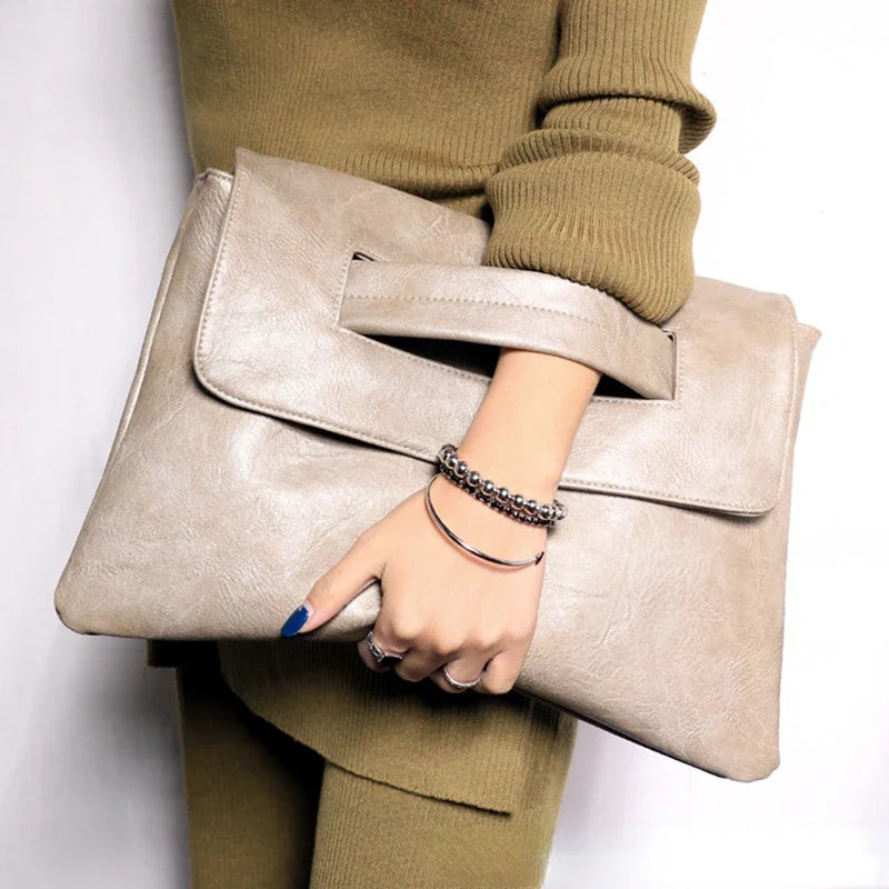 A hand holding a beige leather crossbody clutch bag against a green sweater.