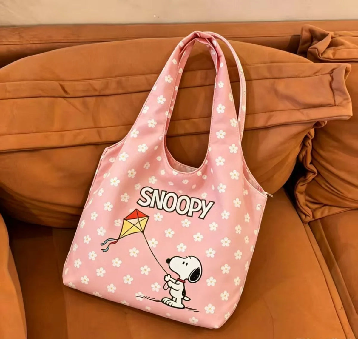 A pink canvas shoulder bag featuring a Snoopy cartoon character resting on a brown couch.