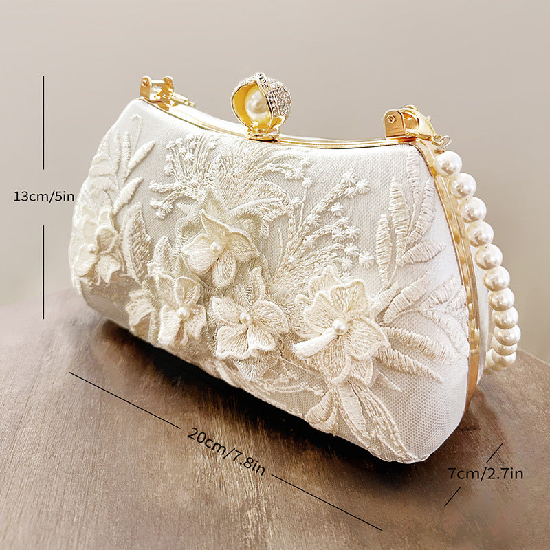 A cream-colored clutch bag with white floral embroidery and a pearl chain strap, shown in profile.