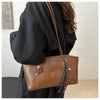 A brown leather tote bag with a dark brown strap is shown carried by a person wearing a black garment.