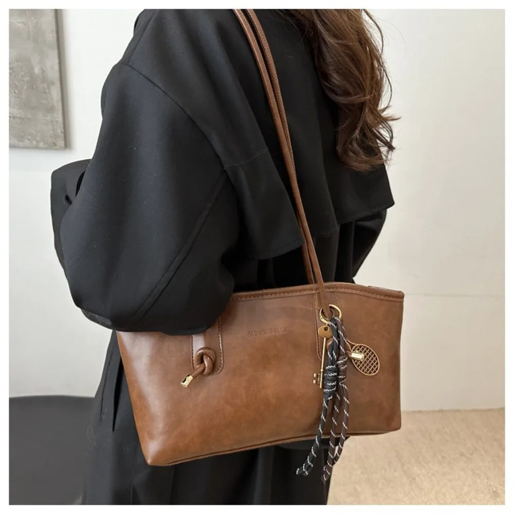 A brown leather tote bag with a dark brown strap is shown carried by a person wearing a black garment.