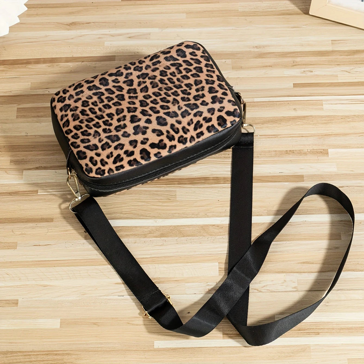 A leopard print crossbody bag with a black strap lying on a light wooden surface.
