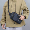 A man wearing a tan jacket holds a navy blue nylon crossbody sling bag.