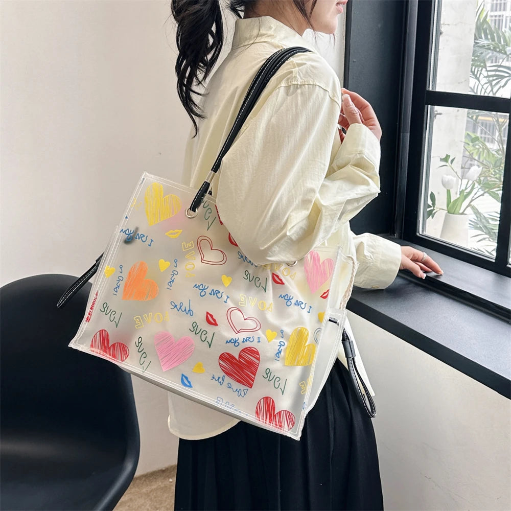 A woman holds a clear plastic shoulder bag decorated with various colored heart shapes and text against a window.