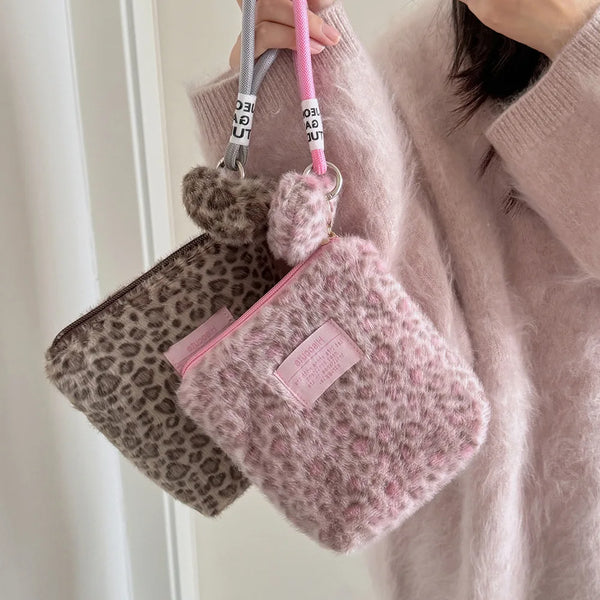 Sac de maquillage à imprimé léopard rose et marron suspendu à une sangle, tenu par une personne vêtue d'un pull rose pâle.