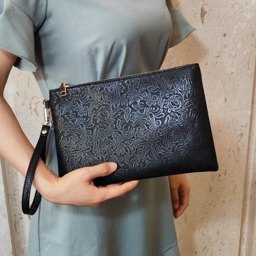 Una mujer sostiene un bolso de cuero sintético negro con estampado floral contra un vestido verde claro.