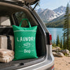 A green laundry bag is placed in the trunk of a silver SUV with a scenic mountain and lake view in the background.