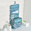 A light blue multi-compartment travel pouch with a floral print hanging on a white tiled wall.