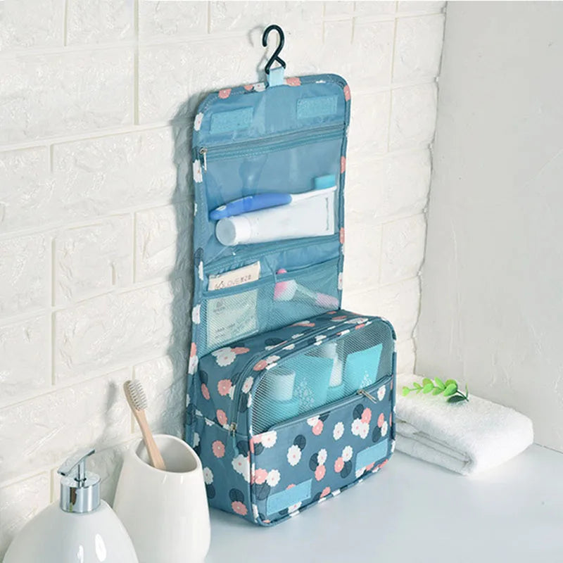 A light blue multi-compartment travel pouch with a floral print hanging on a white tiled wall.