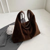 A dark brown suede slouchy shoulder bag resting on a white surface next to a gray blazer.