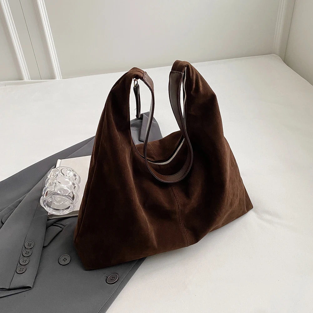 A dark brown suede slouchy shoulder bag resting on a white surface next to a gray blazer.