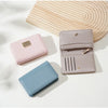 Drei Leder-Kartenwallets in rosa, blau und mauve auf einer hellen Oberfläche neben einem Holz-Hintergrund.
