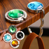 A hand holds several round acrylic foldable hooks in various colors including blue green and amber on a wooden surface.