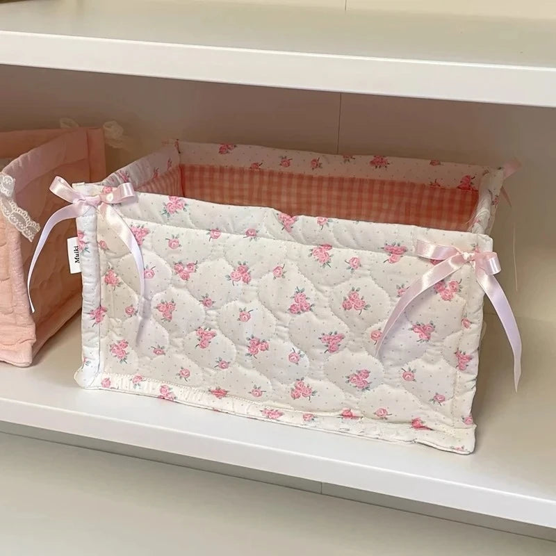 A white quilted cotton storage basket with a pink lace flower design is displayed on a shelf.