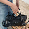 A person holds a black saddle-shaped PU leather city tote bag with black leather straps.