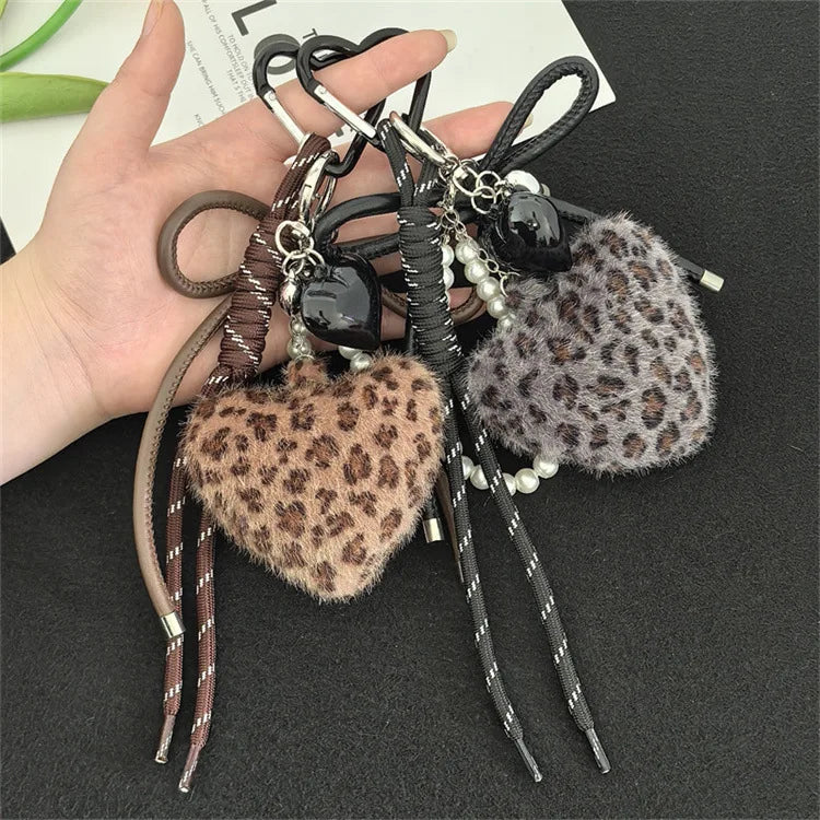 A hand holds a leopard print plush love pendant keychain with black and silver beads and leather cords.