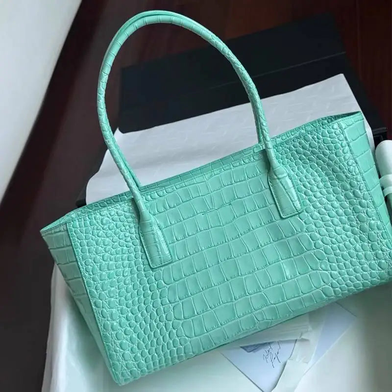 A mint green crocodile print leather handbag with a handle is displayed on a white surface.