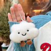 A hand holds a white cloud shaped plush keychain with black eyes and brown ears against a blue knitted background.