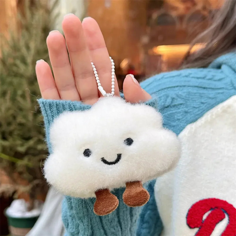 A hand holds a white cloud shaped plush keychain with black eyes and brown ears against a blue knitted background.