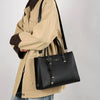 Eine Frau hält eine schwarze, echte Leder-Tasche mit goldenen Beschlägen unter einer tanfarbenen Jacke.