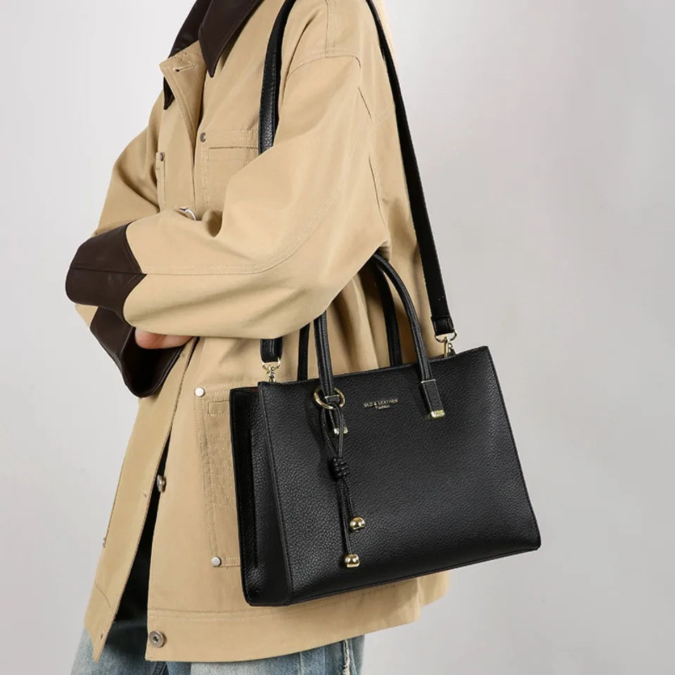 Eine Frau hält eine schwarze, echte Leder-Tasche mit goldenen Beschlägen unter einer tanfarbenen Jacke.