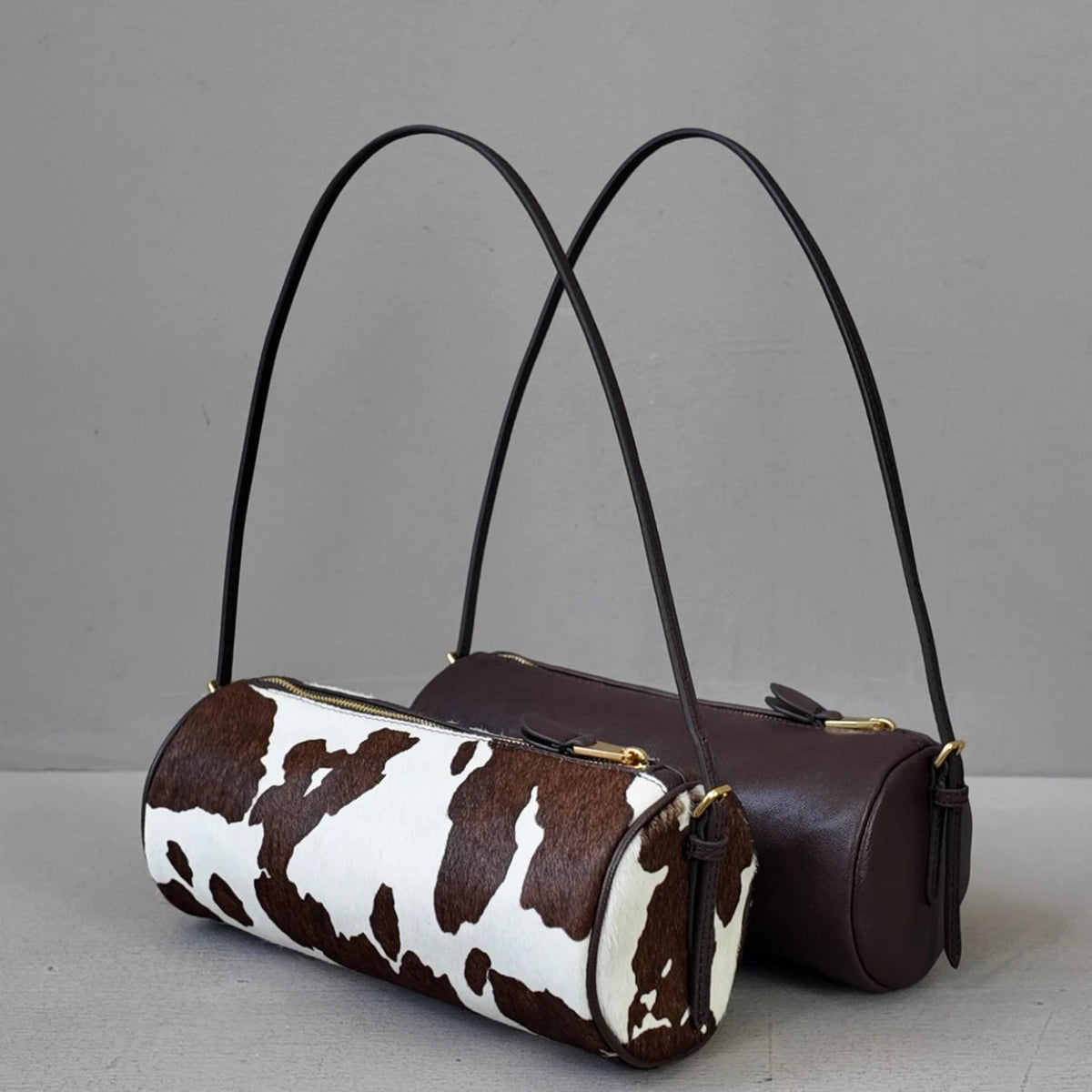 A brown leather cylinder shoulder bag with a white cow print fabric detail.