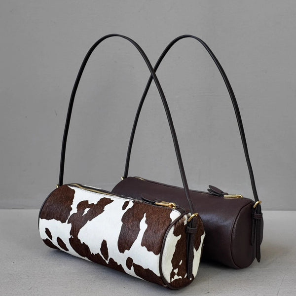 A brown leather cylinder shoulder bag with a white cow print fabric detail.