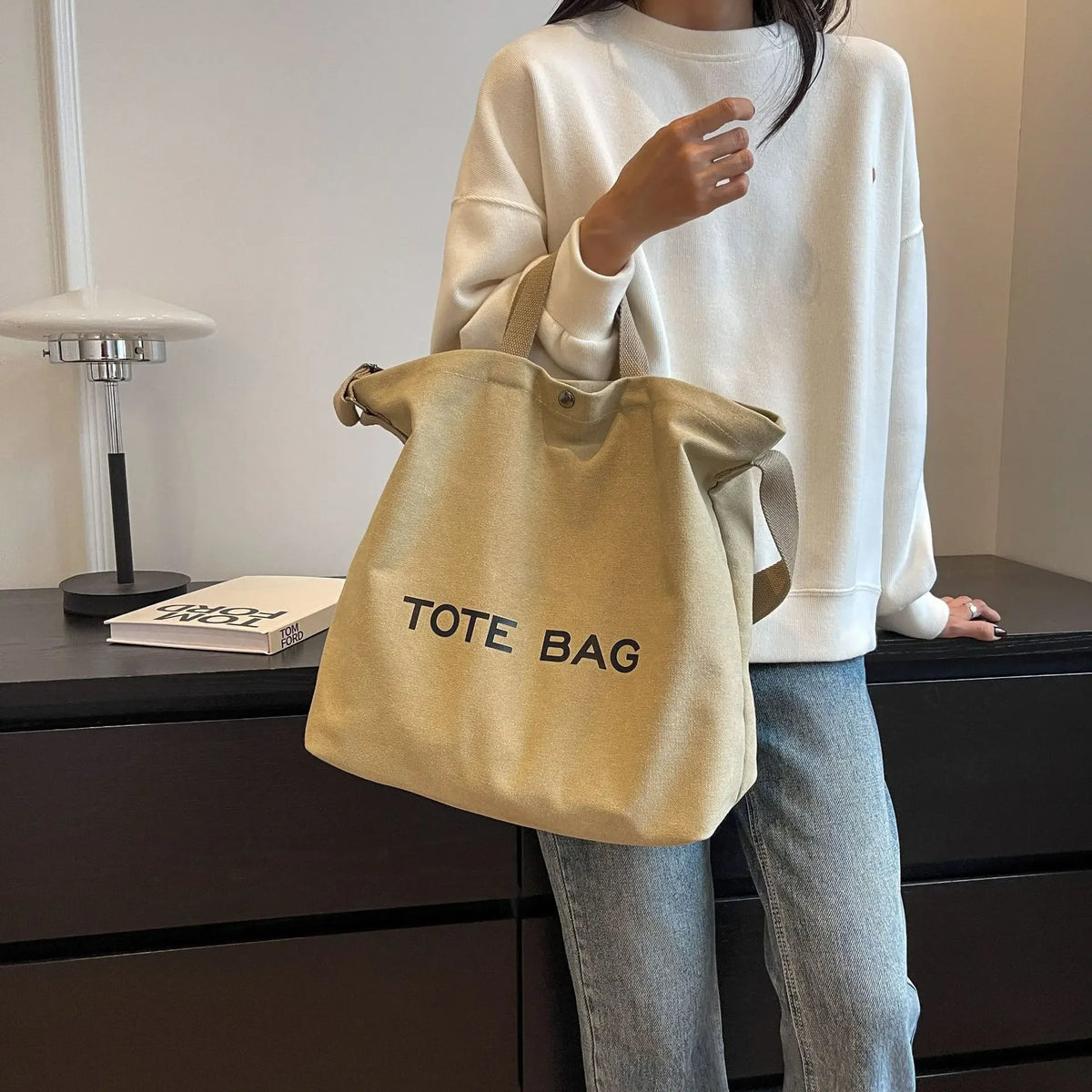 A person is holding a beige canvas tote bag with the text TOTE BAG in front of a dark cabinet.
