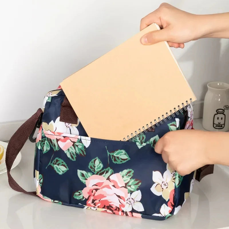 Una mano sostiene un cuaderno beige dentro de una bolsa de almuerzo floral azul marino.