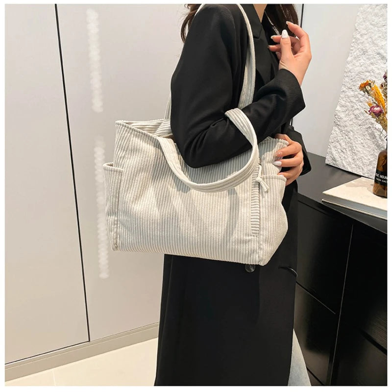 A woman holds a light gray corduroy casual handbag against a dark background.
