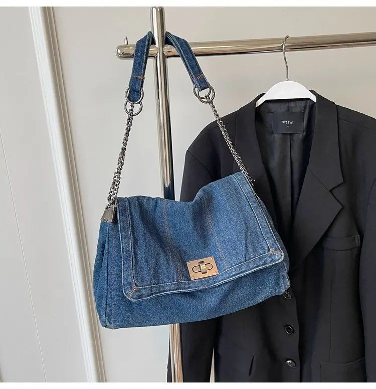 A dark blue denim tote bag with a chain strap is displayed hanging on a hanger against a light background.
