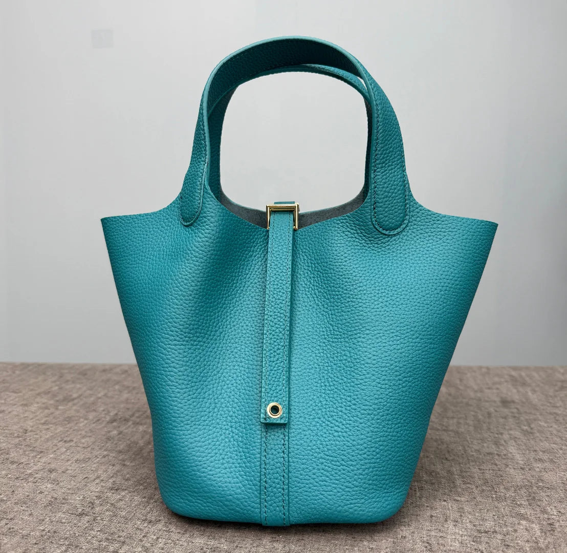 A teal genuine leather bucket bag with a gold-toned clasp and leather trim, shown from the front.