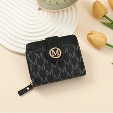Black compact wallet with gold monogram print resting on a light background next to a yellow tulip.