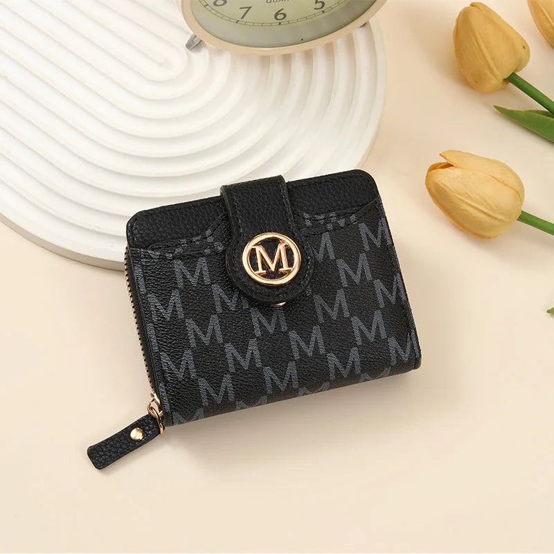 Black compact wallet with gold monogram print resting on a light background next to a yellow tulip.