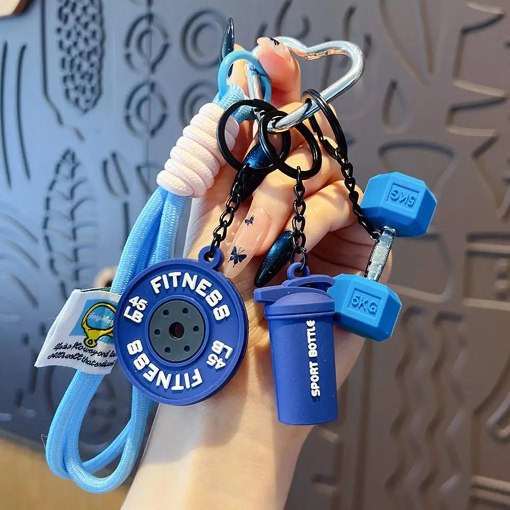 A hand holds a fitness keychain featuring a dumbbell and kettlebell charm attached to a blue cord.