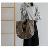 A woman holds a leopard print canvas crossbody tote bag with brown leather straps against a light gray sweater and black skir